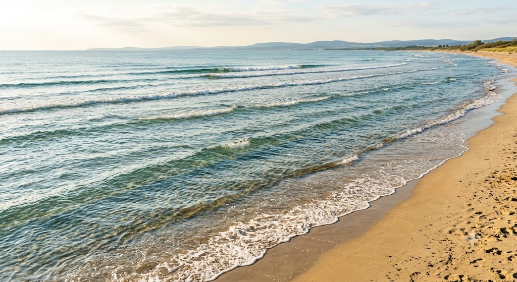 Landschaftsaufnahme eines Sandstrands mit vielen flachen, gleichmäßigen Meereswellen, die nacheinander ans Ufer rollen.